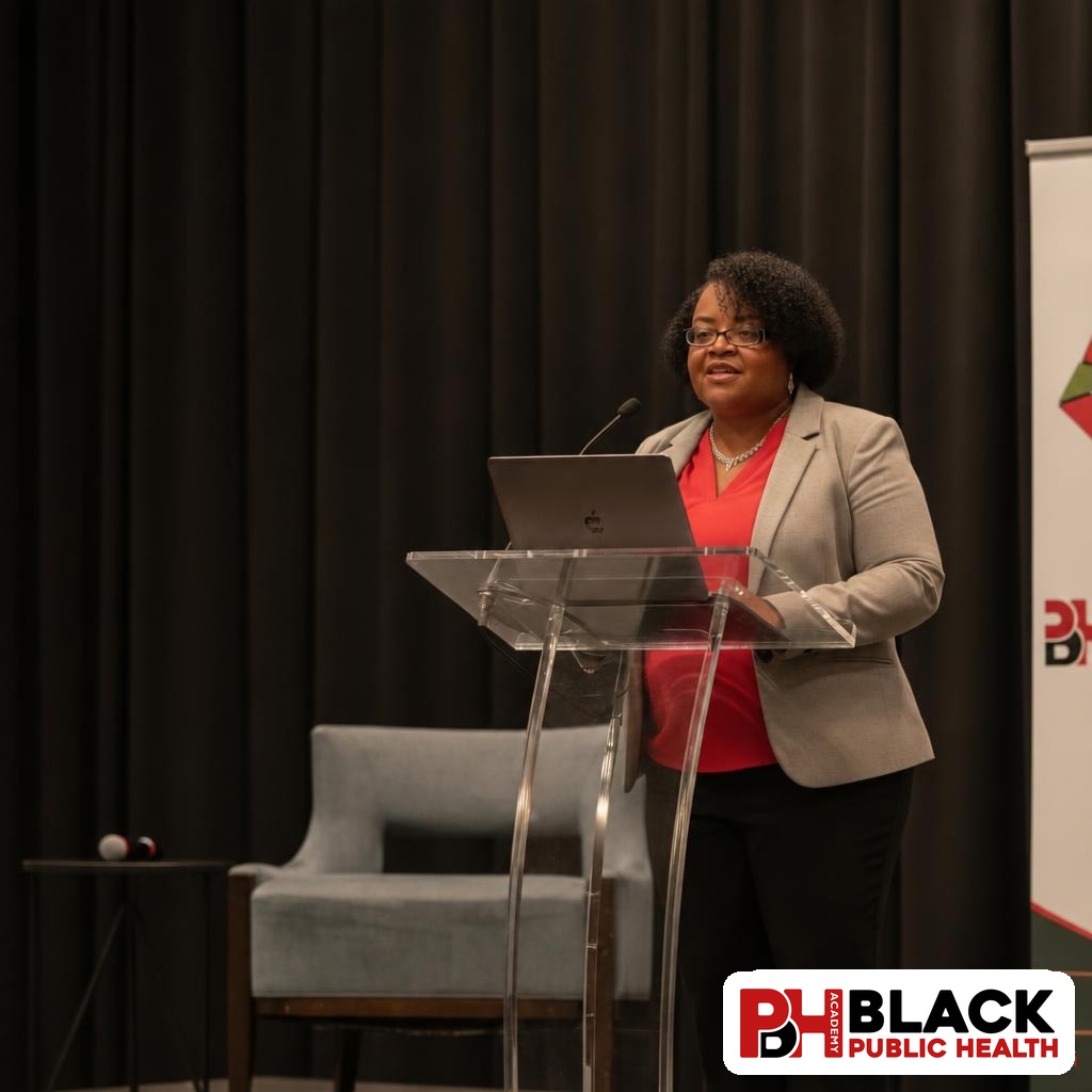Leisha McKinley-Beach speaking at a clear lectern with a laptop, wearing a gray blazer and coral blouse.